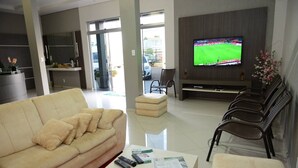 A modern living room with a beige sofa, a coffee table with remote controls and a newspaper, a TV displaying a sports event, and a seating area with chairs. - Hotel Querência (Pitanga)