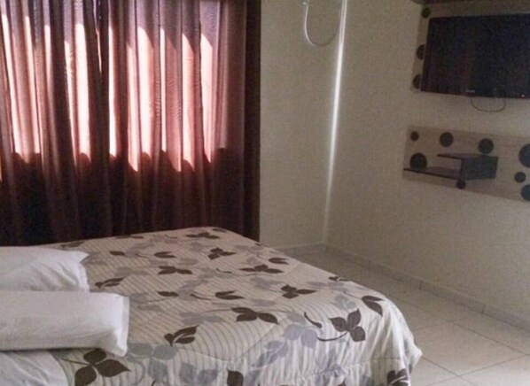 A hotel room with a bed, curtains, a mounted television, and a wall-mounted shelf.