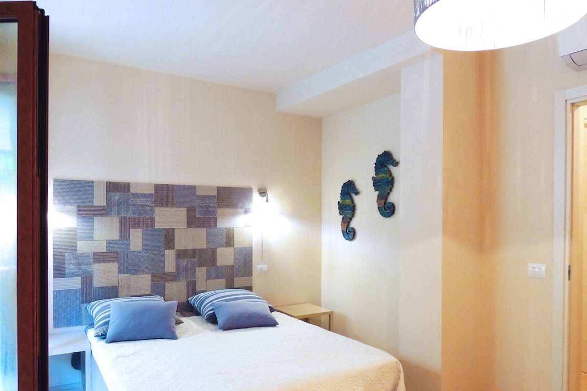 A bedroom with a bed, two nightstands, a wall-mounted light, and two decorative seahorse wall art pieces.