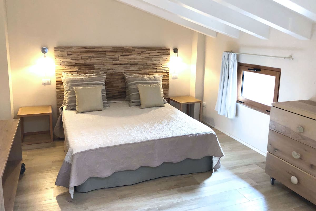A bedroom with a stone headboard, a large bed with multiple pillows, bedside tables, a wooden dresser, and a window with curtains.