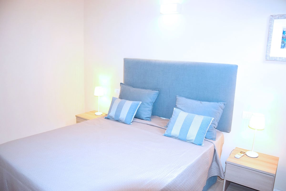 A neatly made bed with a blue headboard and matching pillows, two bedside lamps, and a nightstand with a phone and glasses.