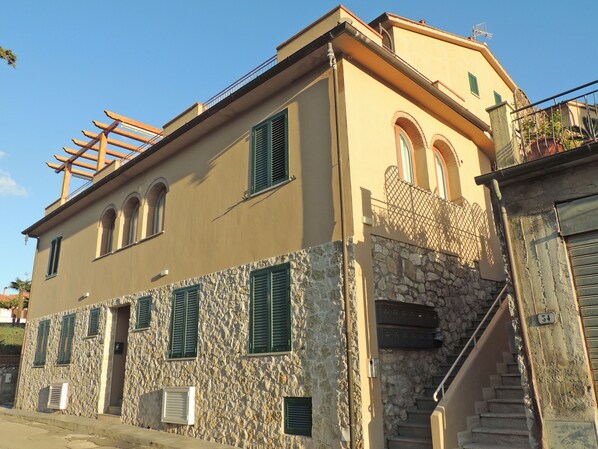 A two-story building with a stone facade, green shutters, and a wooden pergola on the roof.