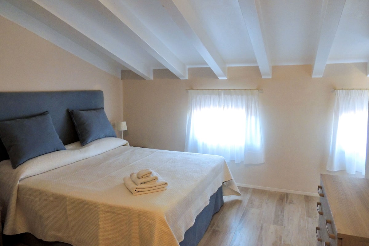 A bedroom with a bed, pillows, a nightstand, a window with curtains, and a wooden floor.