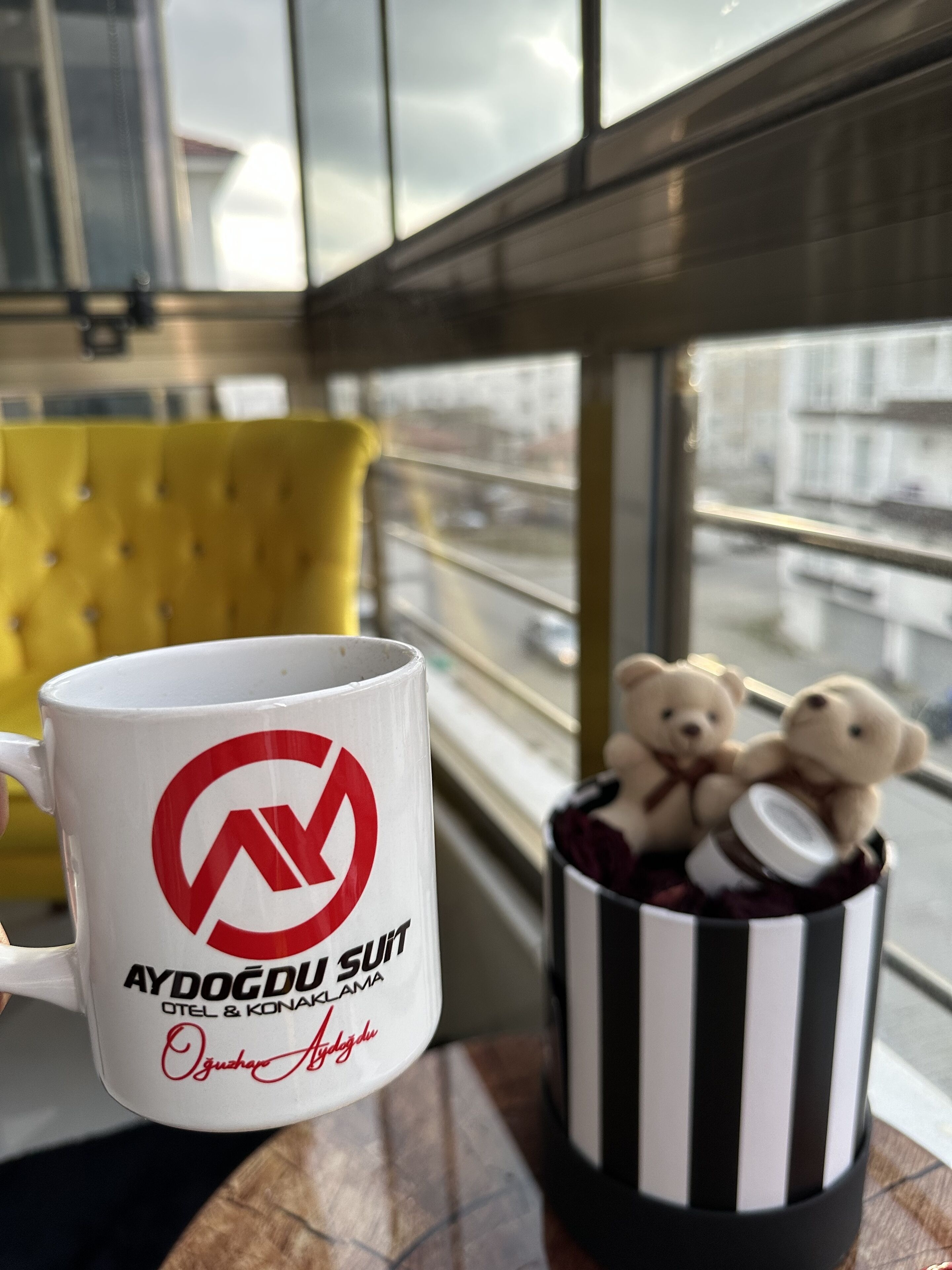 Une tasse blanche ornée d’un logo et d’un texte, une chaise jaune et une tasse rayée noire et blanche avec des motifs d’ours en peluche.