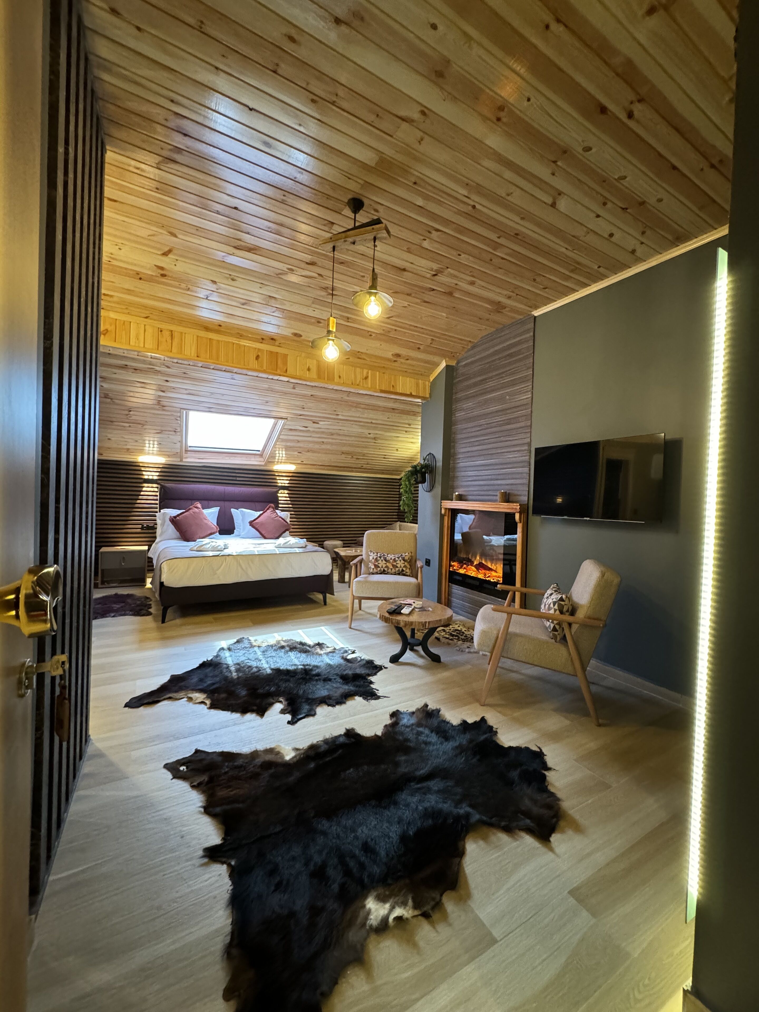A modern bedroom with a wooden ceiling, a bed with pillows, a fireplace, and leather rugs.