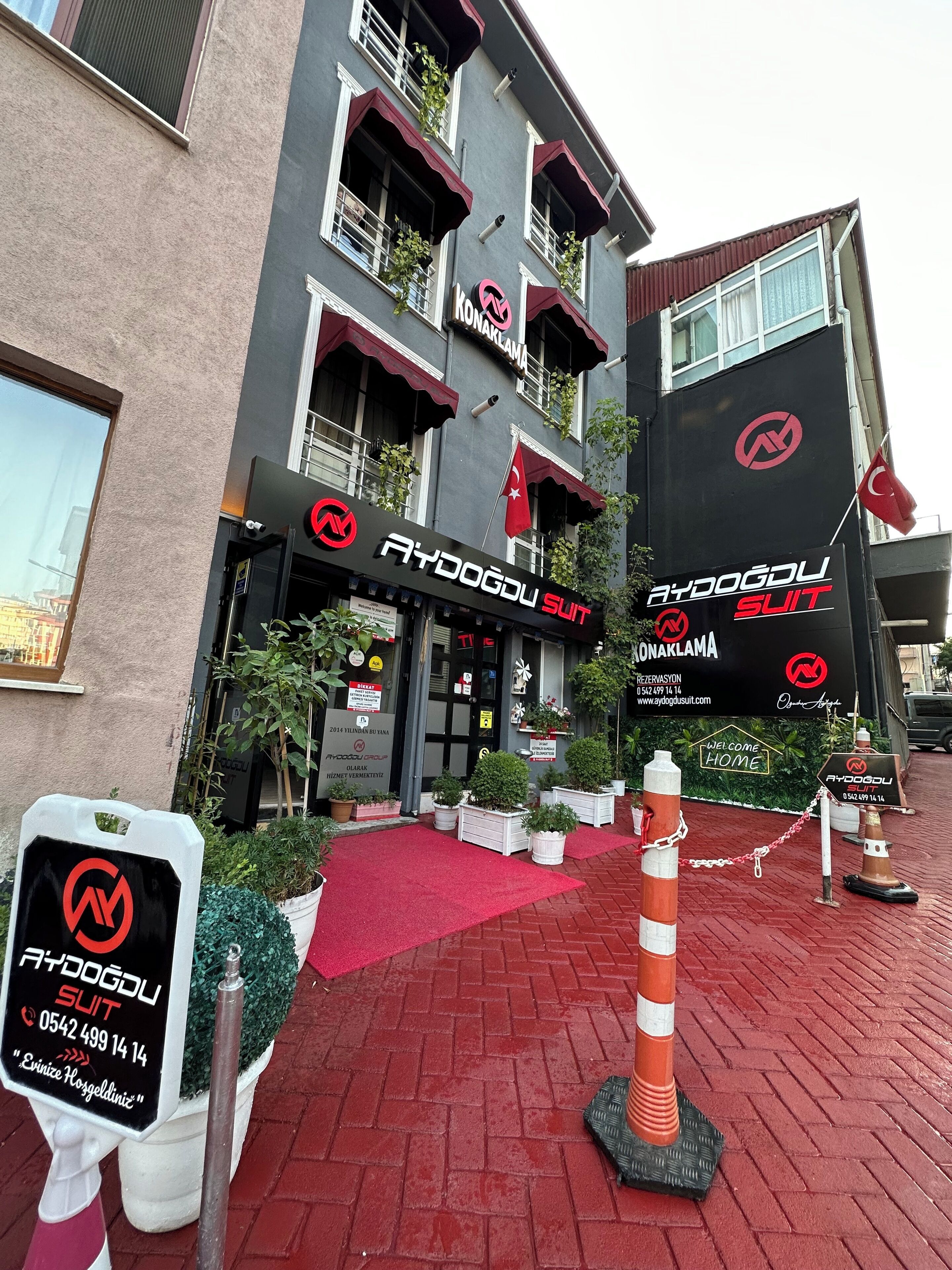 A building with multiple entrances, each marked with a sign for 'Aydogdu Suit'. There are potted plants and a red carpet leading to the entrance.