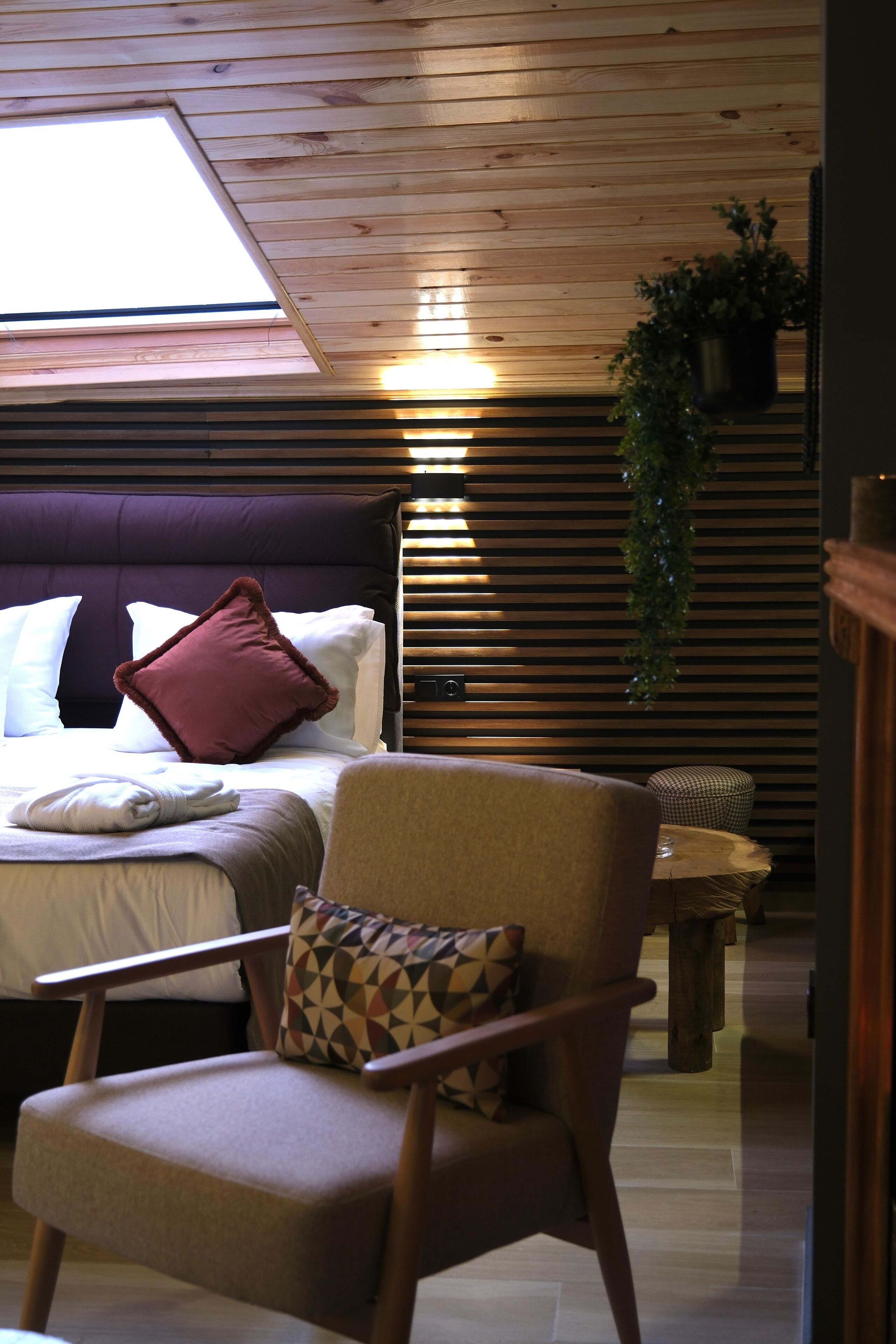 Ein Schlafzimmer mit Bett, Stuhl, Nachttisch, einer hängenden Pflanze und einem Dachfenster.