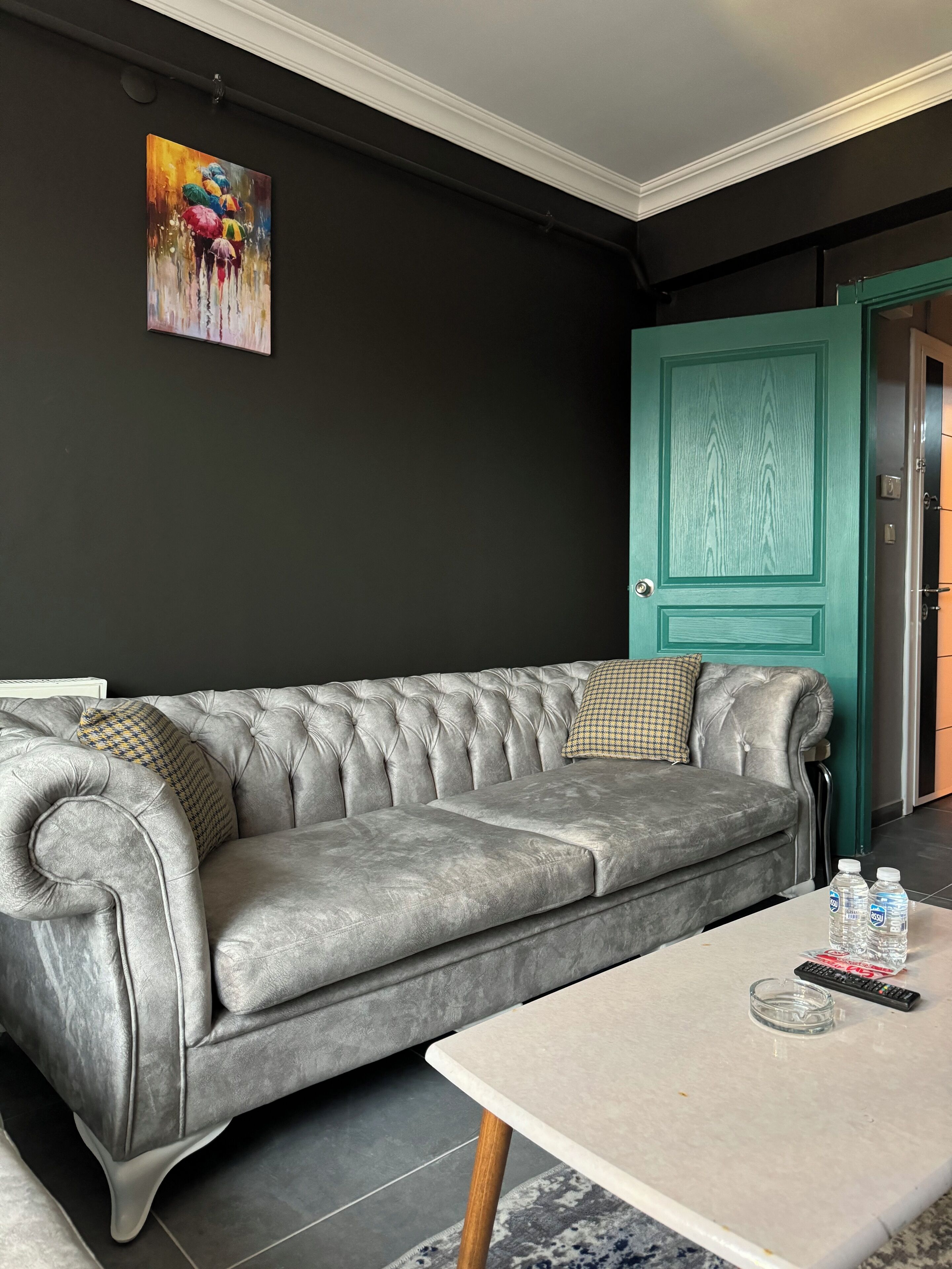 Un sofá gris con tapicería texturizada, cojines estampados, una mesa de centro de mármol con controles remotos y botellas de agua, y una puerta color turquesa al fondo.
