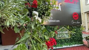 A sign with the text "DOGSUIT" and a website address, surrounded by greenery and a "WELCOME HOME" sign.