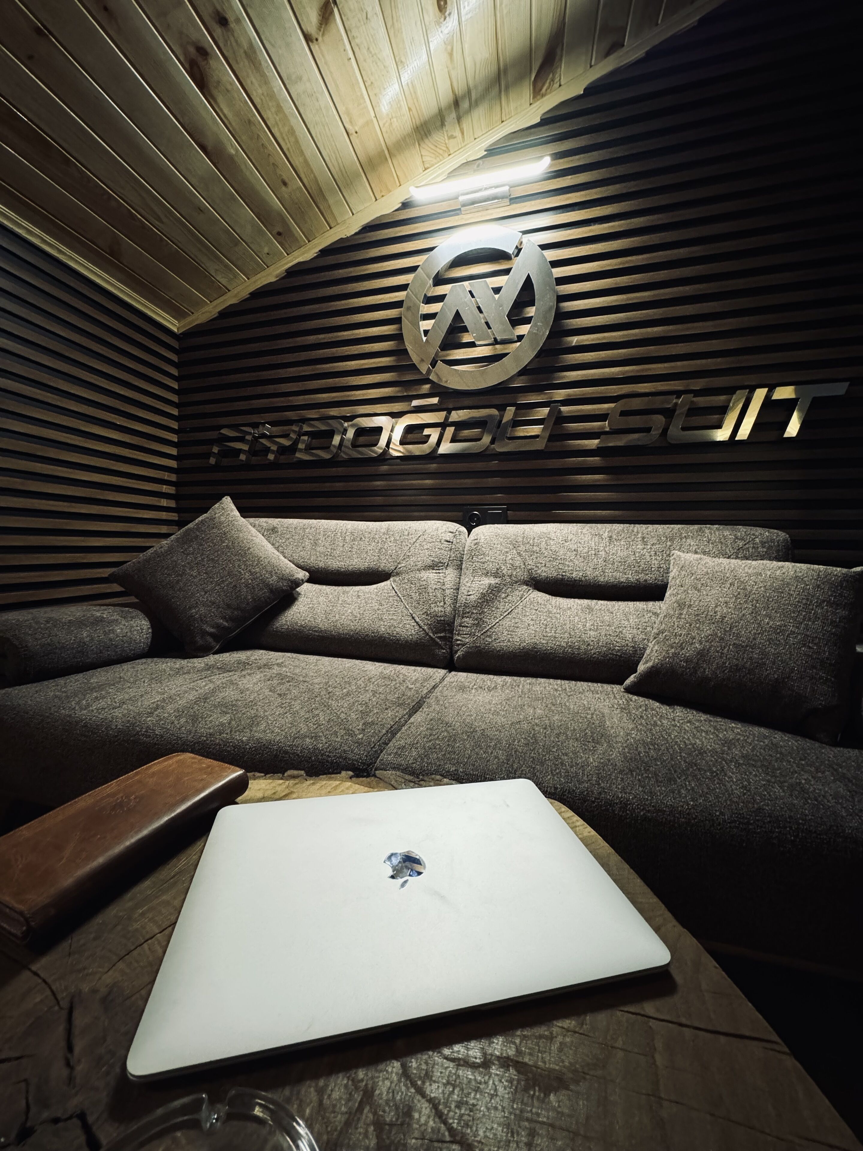 A modern room with a wooden slatted ceiling, a grey sofa with cushions, and a laptop on a wooden table.