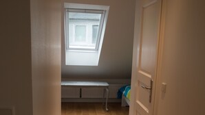 Un couloir étroit, doté d’un plancher en bois, mène à une pièce dont le plafond est en pente et qui possède une fenêtre.