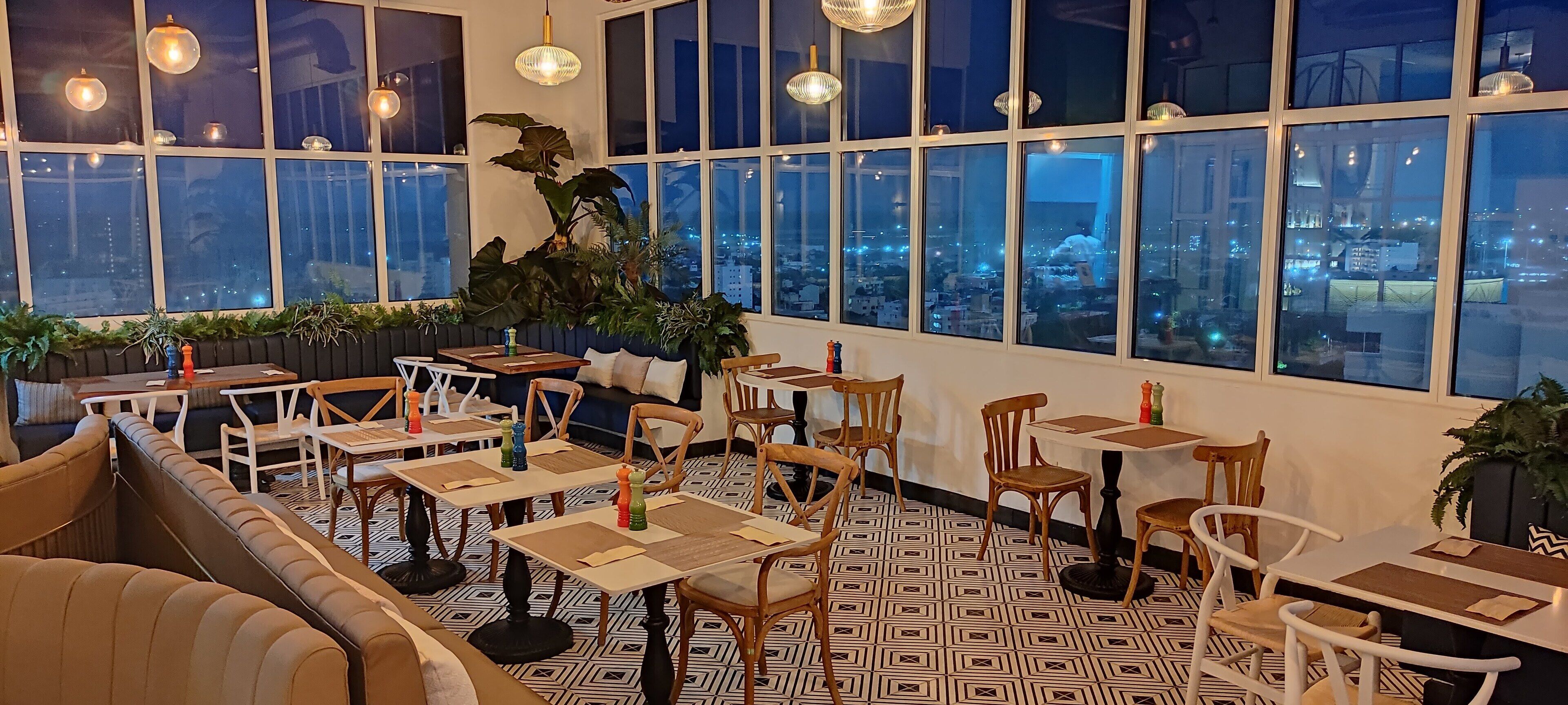 Un restaurante con varias mesas y sillas, amplios ventanales con vista a la ciudad y lámparas colgantes.