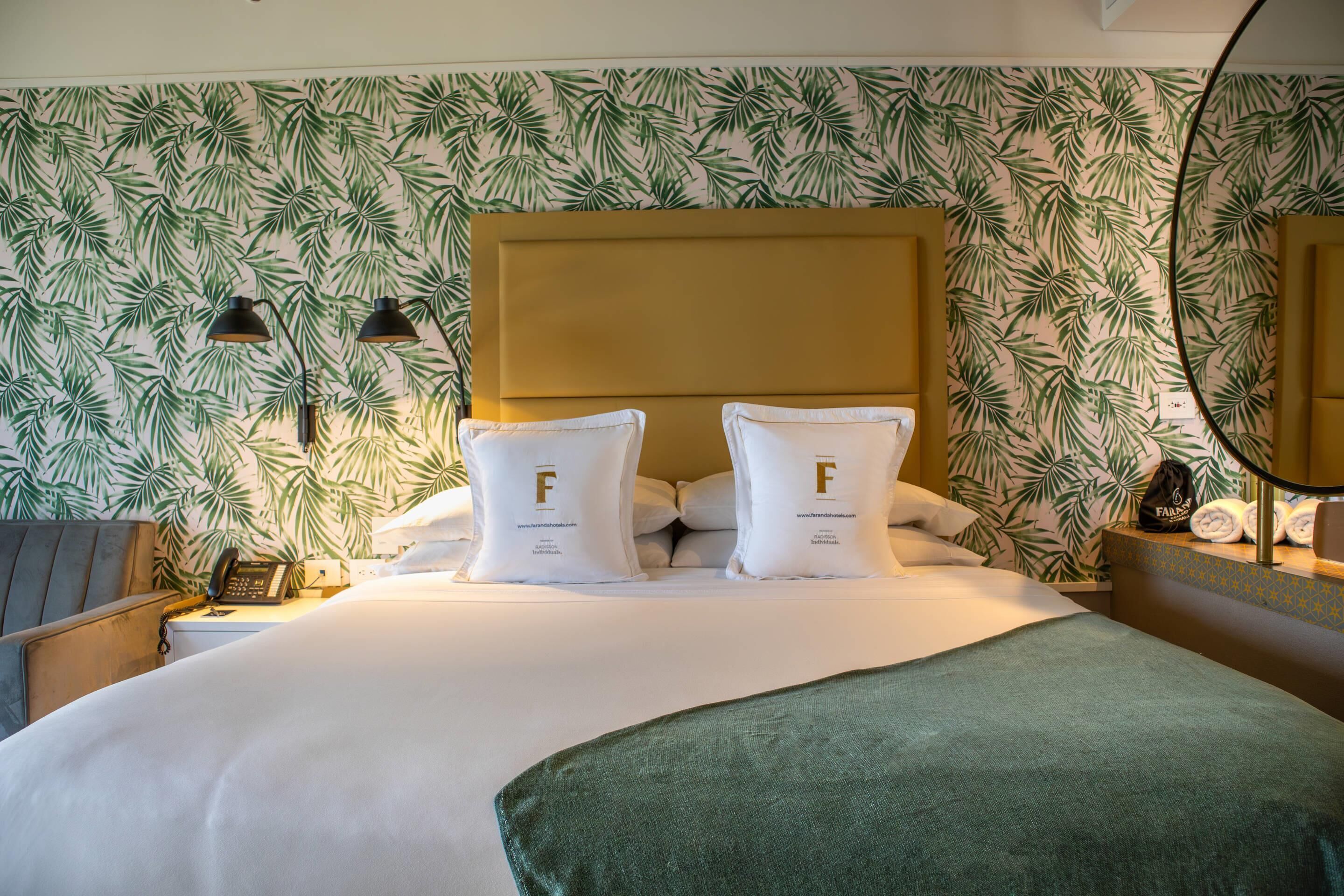 A hotel room with a bed, two pillows with a logo, a nightstand with a lamp, a telephone, and a wall with a leaf pattern.