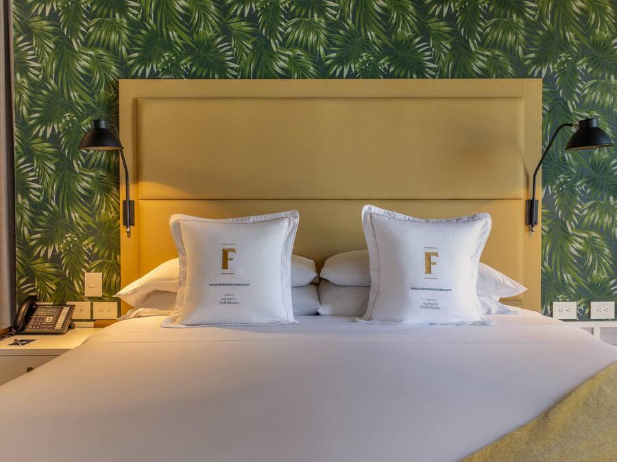 A bed with a headboard, two pillows with a logo, a bedside lamp, a phone, and a wall with a leaf pattern.