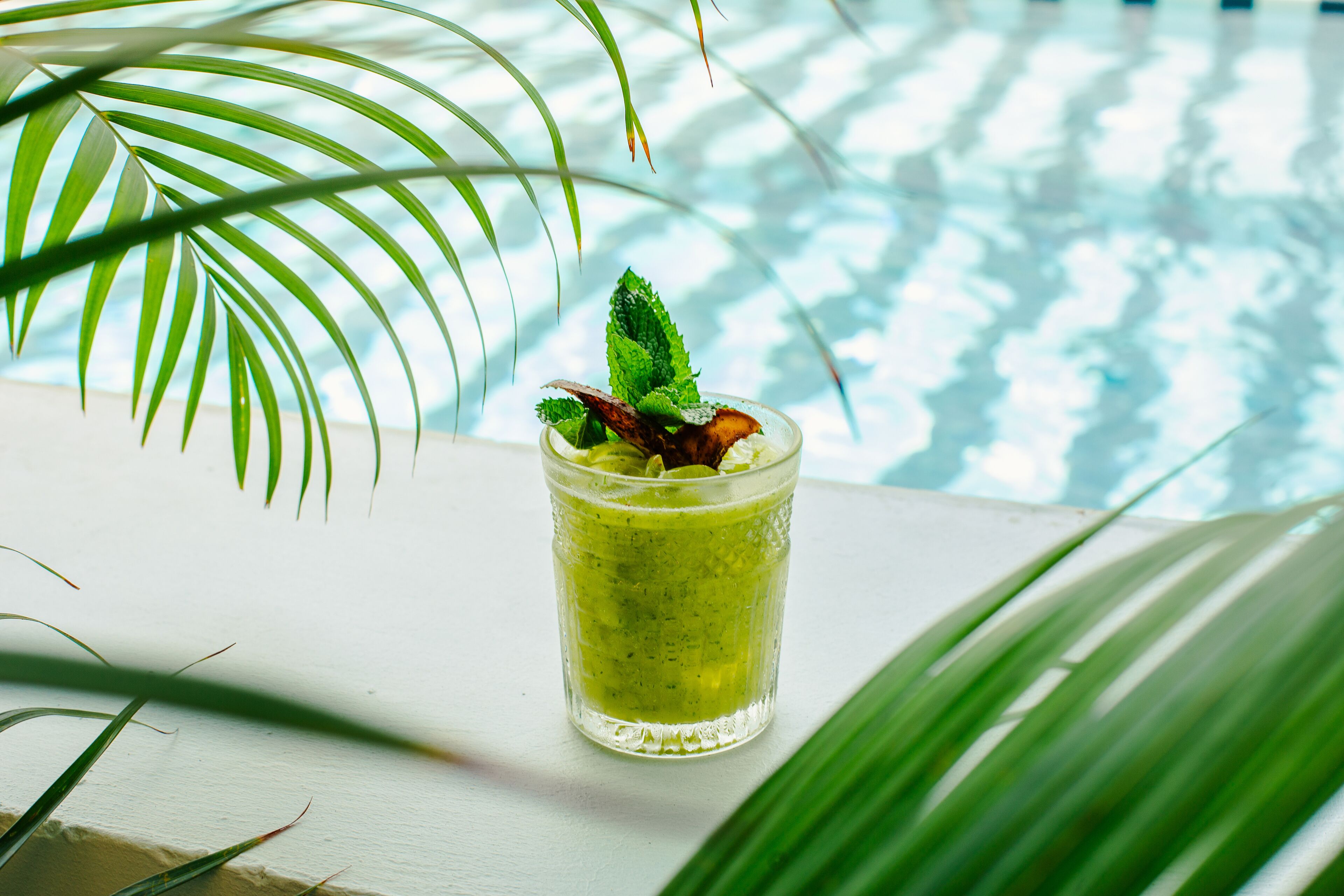 A glass of green beverage with mint leaves, placed on a white surface with a pool in the background.