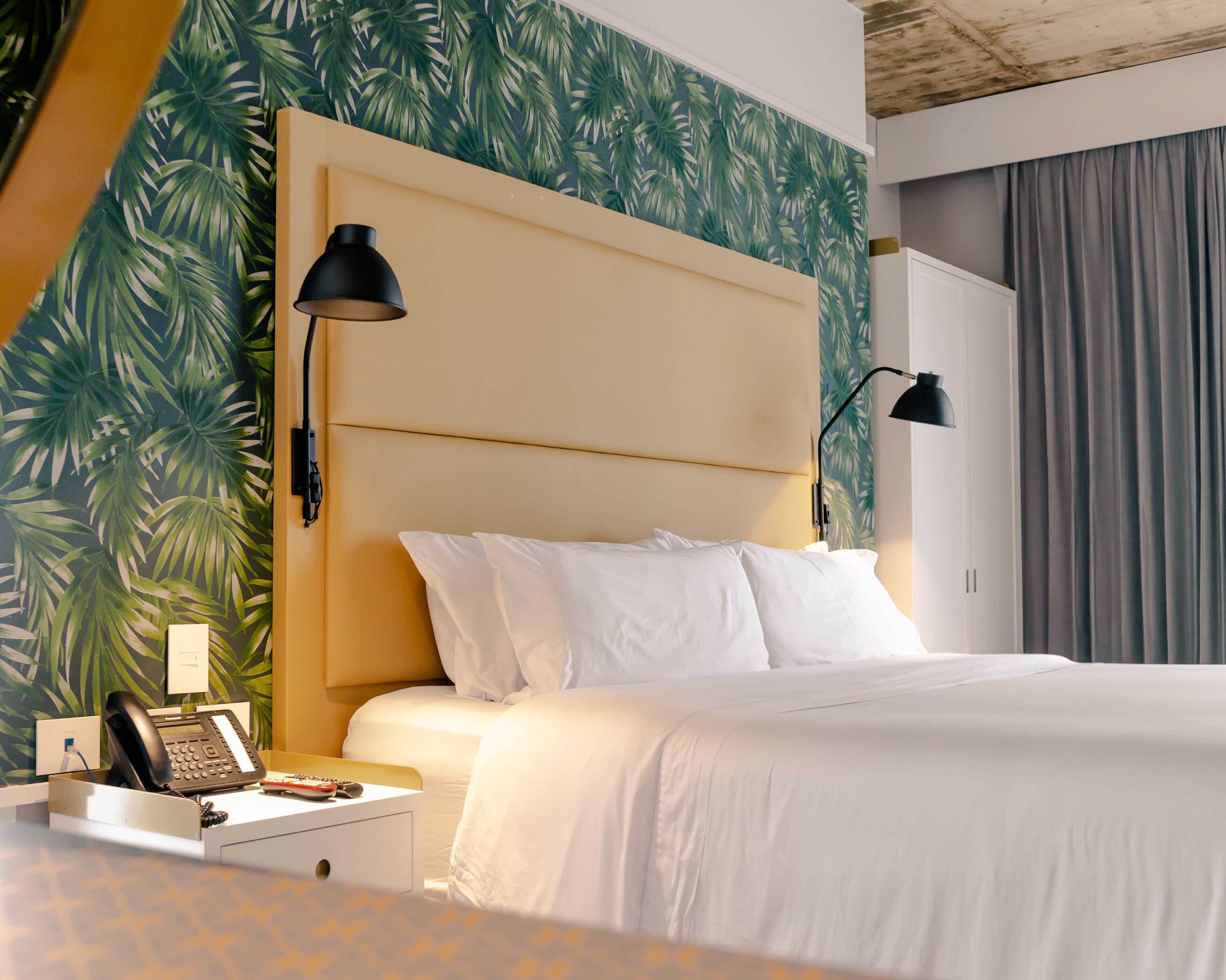 Habitación de hotel con cama, lámparas de noche, mesita de noche con teléfono y papel tapiz con hojas tropicales.