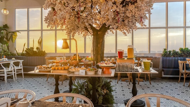 Un espace repas agrémenté d’un grand arbre servant de décoration centrale, de tables garnies de nourriture et offrant une vue sur la ville grâce à de grandes fenêtres.