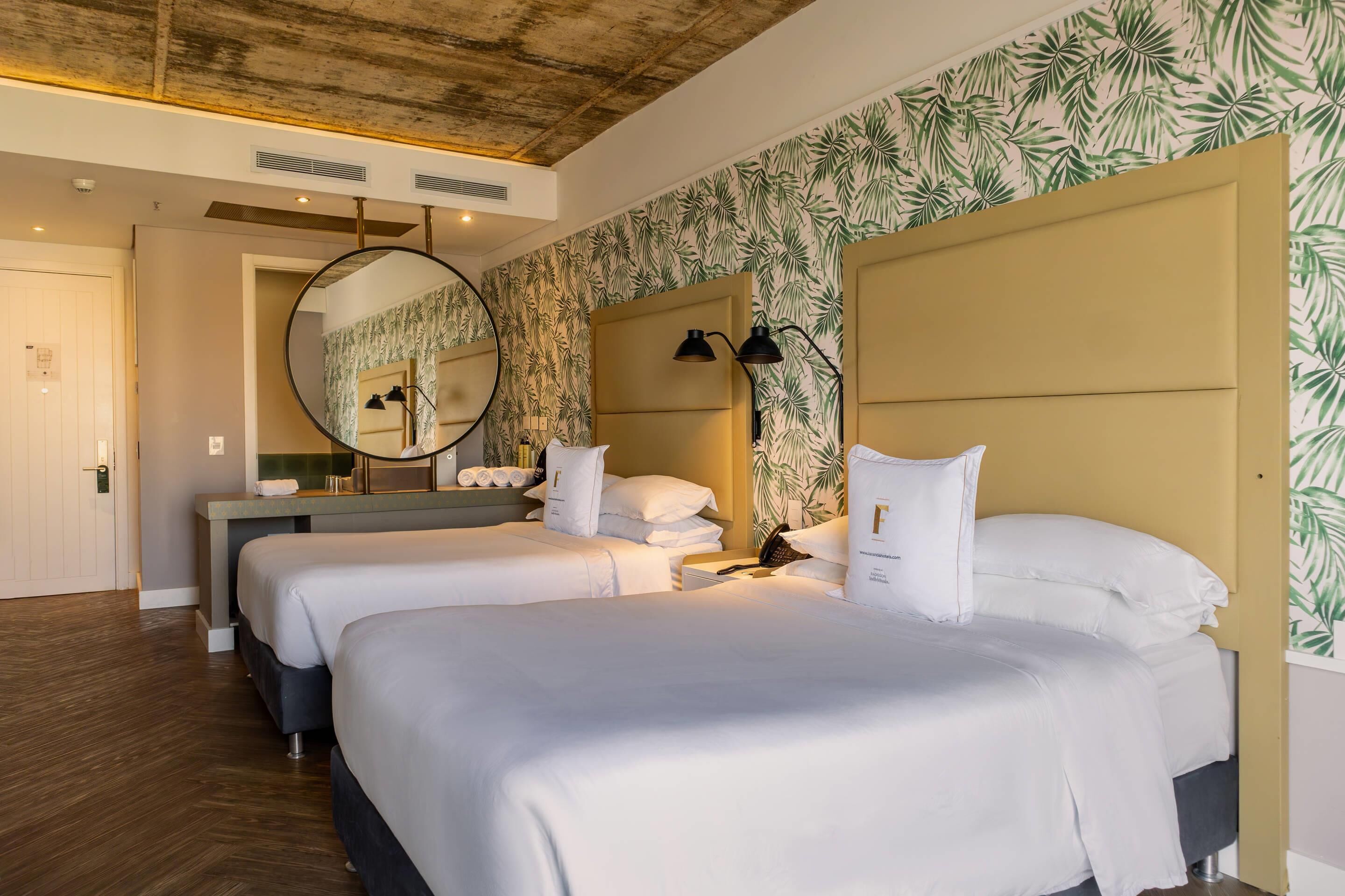Habitación de hotel con dos camas, un espejo redondo, una mesita de noche con lámpara y una pared con diseño de hojas.