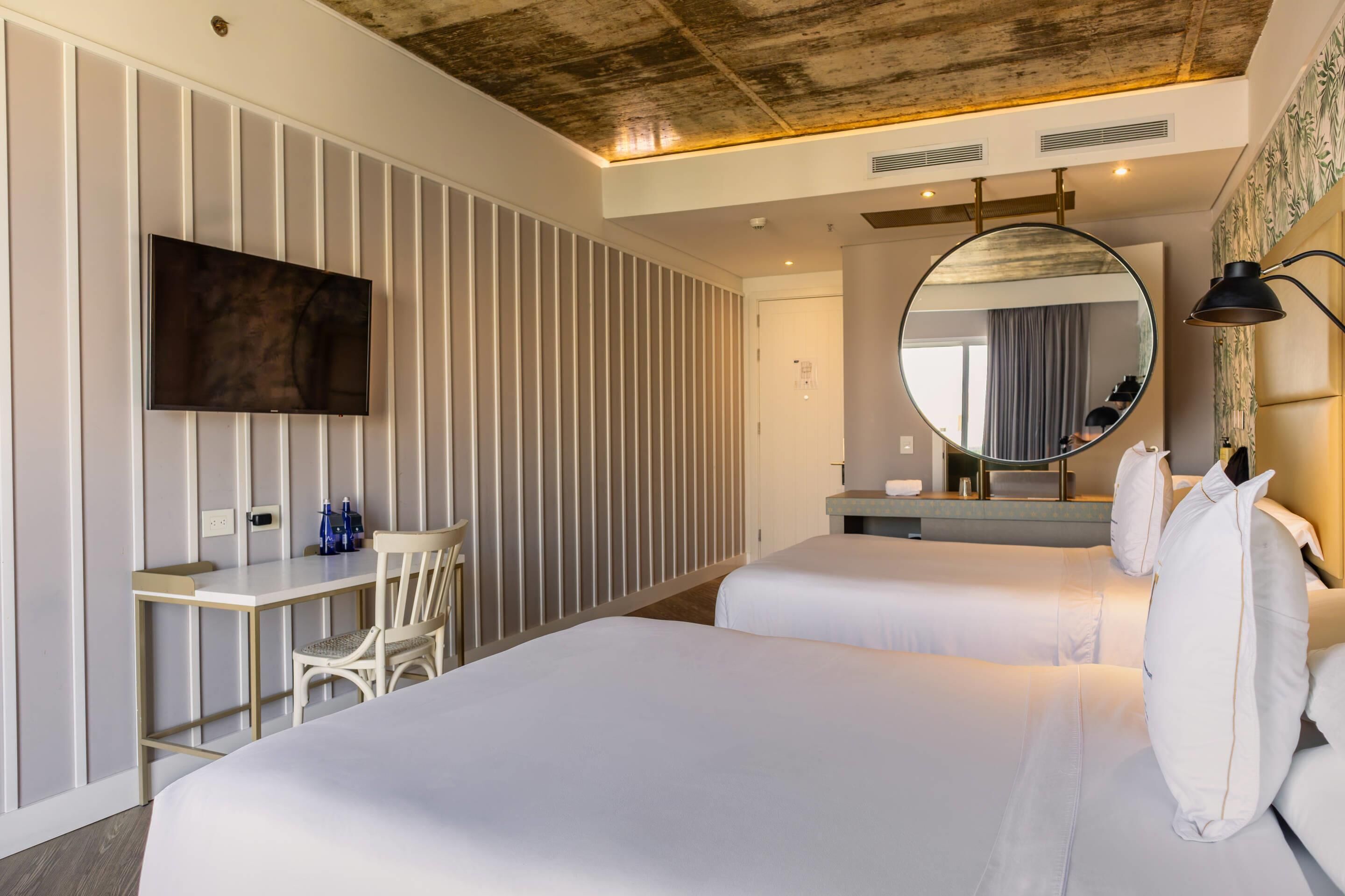 A hotel room with two beds, a desk, a chair, a TV, a mirror, and a lamp.