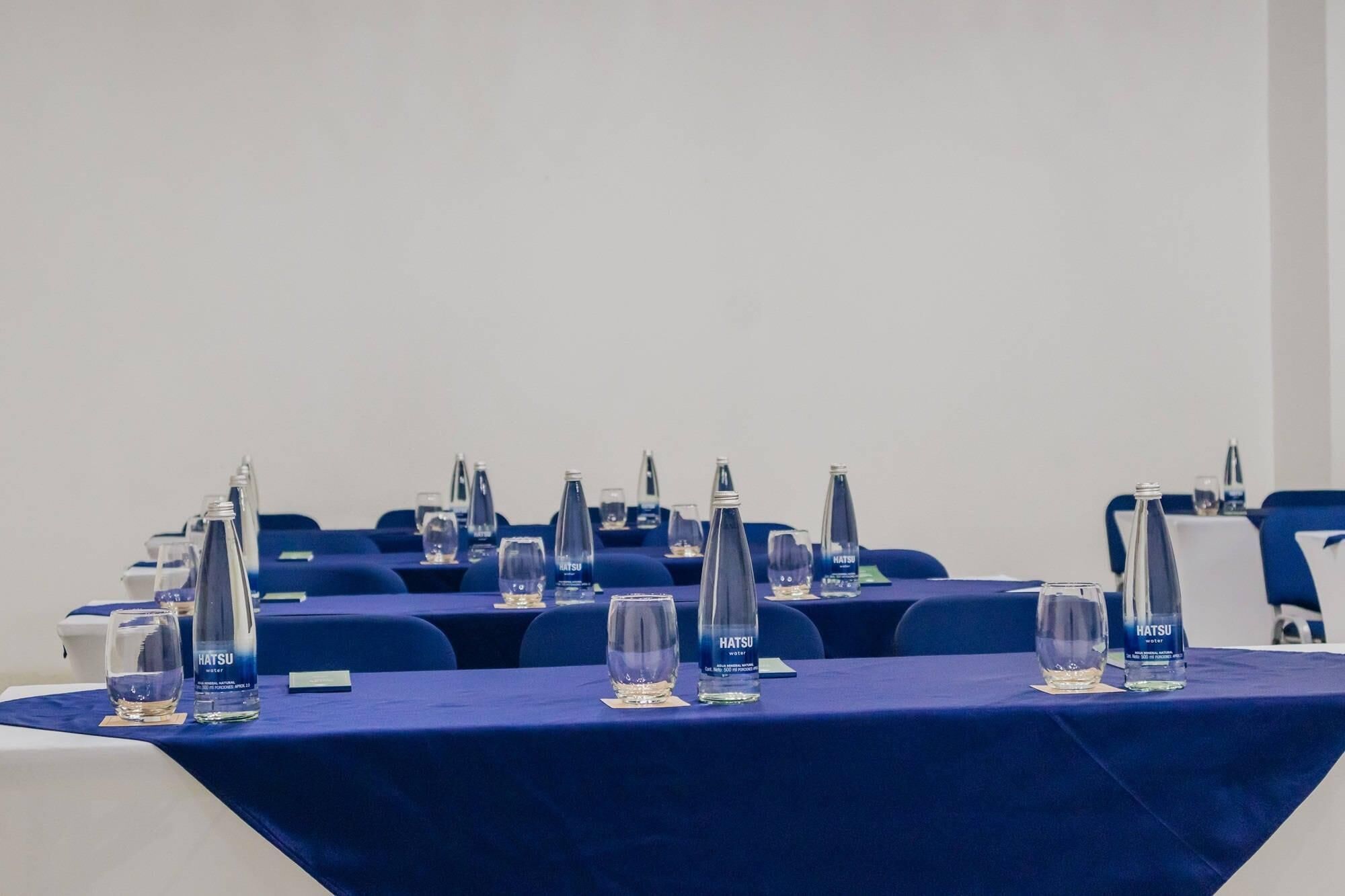Une salle de conférence avec des tables préparées pour une réunion, chacune étant équipée d’une bouteille d’eau et d’un verre.