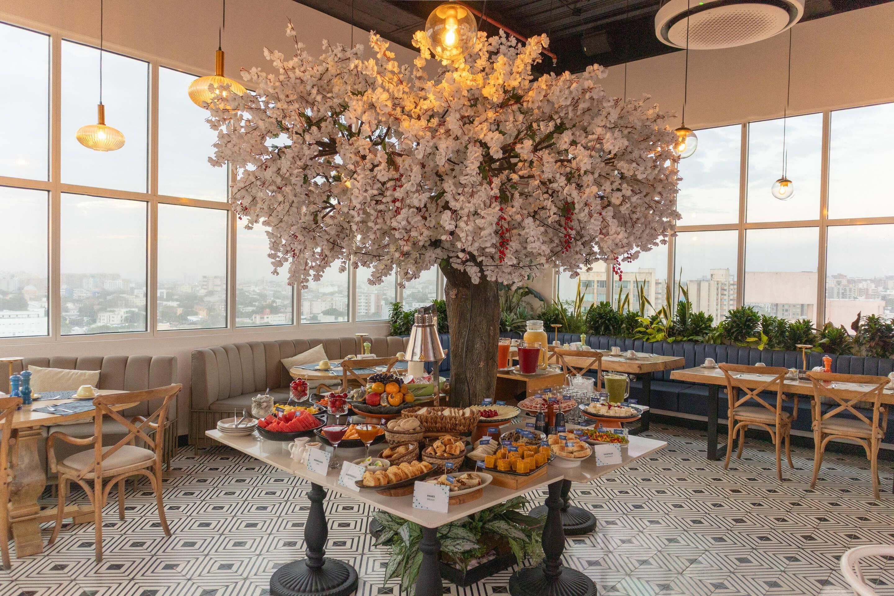 A dining area with a large tree centerpiece, a buffet table, and a city view through large windows.