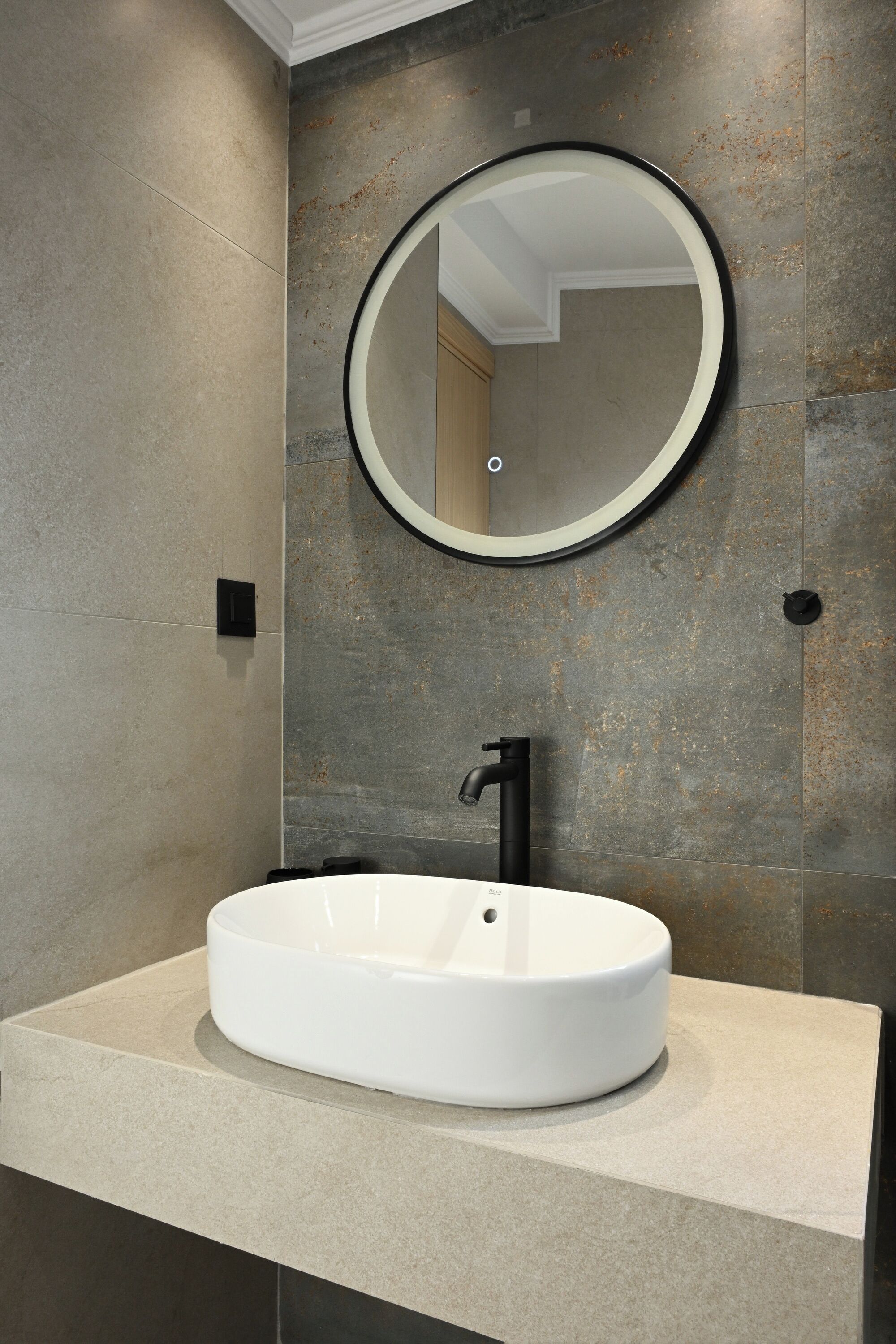 Un baño moderno con un lavamanos ovalado grande, un espejo redondo y un grifo negro.