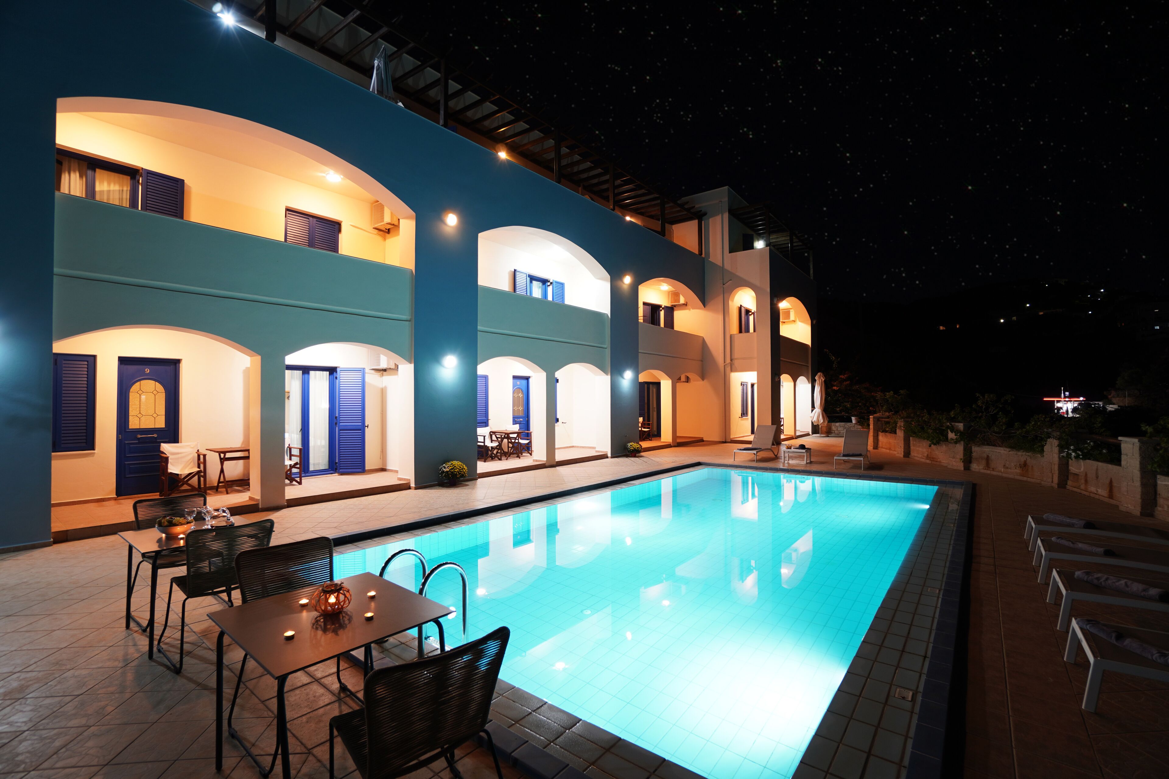 A nighttime swimming pool area with a two-story building, outdoor seating, and a clear view of the pool.