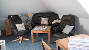 Een knus slaapkamerloft met een schuine plafond, een houten bijzettafeltje, twee leren fauteuils, een klein houten tafeltje en een enkele roos op de vloer.
