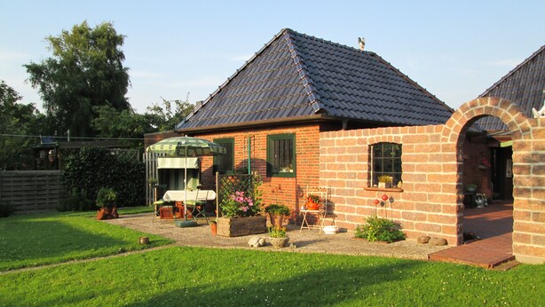 Een bakstenen huis met pannendak, een terras met tafel en stoelen, en een tuin.