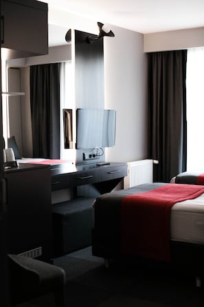 Ein Hotelzimmer mit Schreibtisch, Stuhl, Bett, Fernseher und einem Fenster mit Vorhängen.