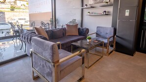 A modern hotel room with a sofa, armchair, and a small table. There is a refrigerator and a window with a view of the city. - Methis Hotel (Izmir)