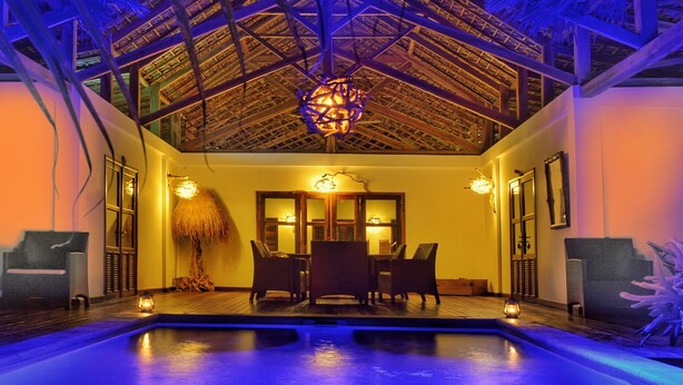 A pool area with a thatched roof, wooden deck, and a dining table with chairs.