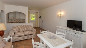 Una sala de estar acogedora con un sofá a cuadros, una mesa de comedor con sillas, un televisor y una zona de cocina con armarios y un ventanal.