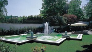 Un jardín ajardinado con una fuente central, rodeado por un seto bajo y una zona pavimentada.