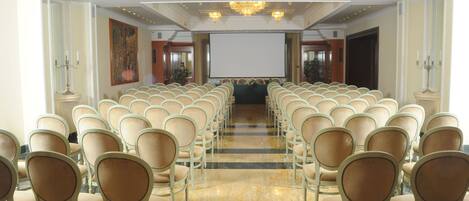 Une salle de conférence avec des rangées de chaises beiges, un grand écran à l’avant et des lustres au plafond.