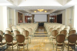 Un salón de conferencias con filas de sillas color beige, una pantalla grande al frente y candelabros en el techo.