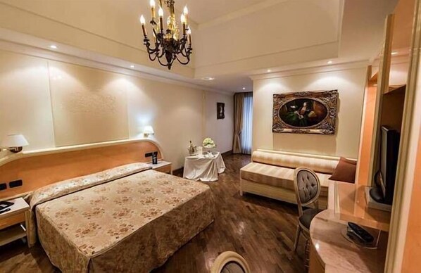 Habitación de hotel con cama doble, sofá, mesita, televisor y candelabro.