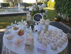 A table set up outdoors with various food items and gift boxes, surrounded by a fountain and greenery. - LH Domus Caesari (Marino)