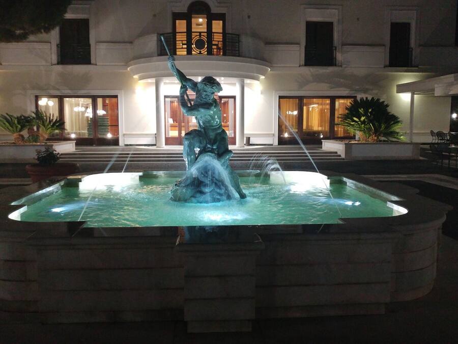A fountain with a statue in the center, surrounded by a pool, in front of a building with multiple windows and steps leading up to the entrance.