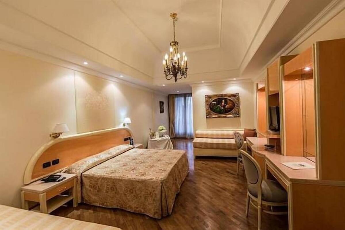 A hotel room with a bed, a desk, a chair, a TV, and a chandelier.