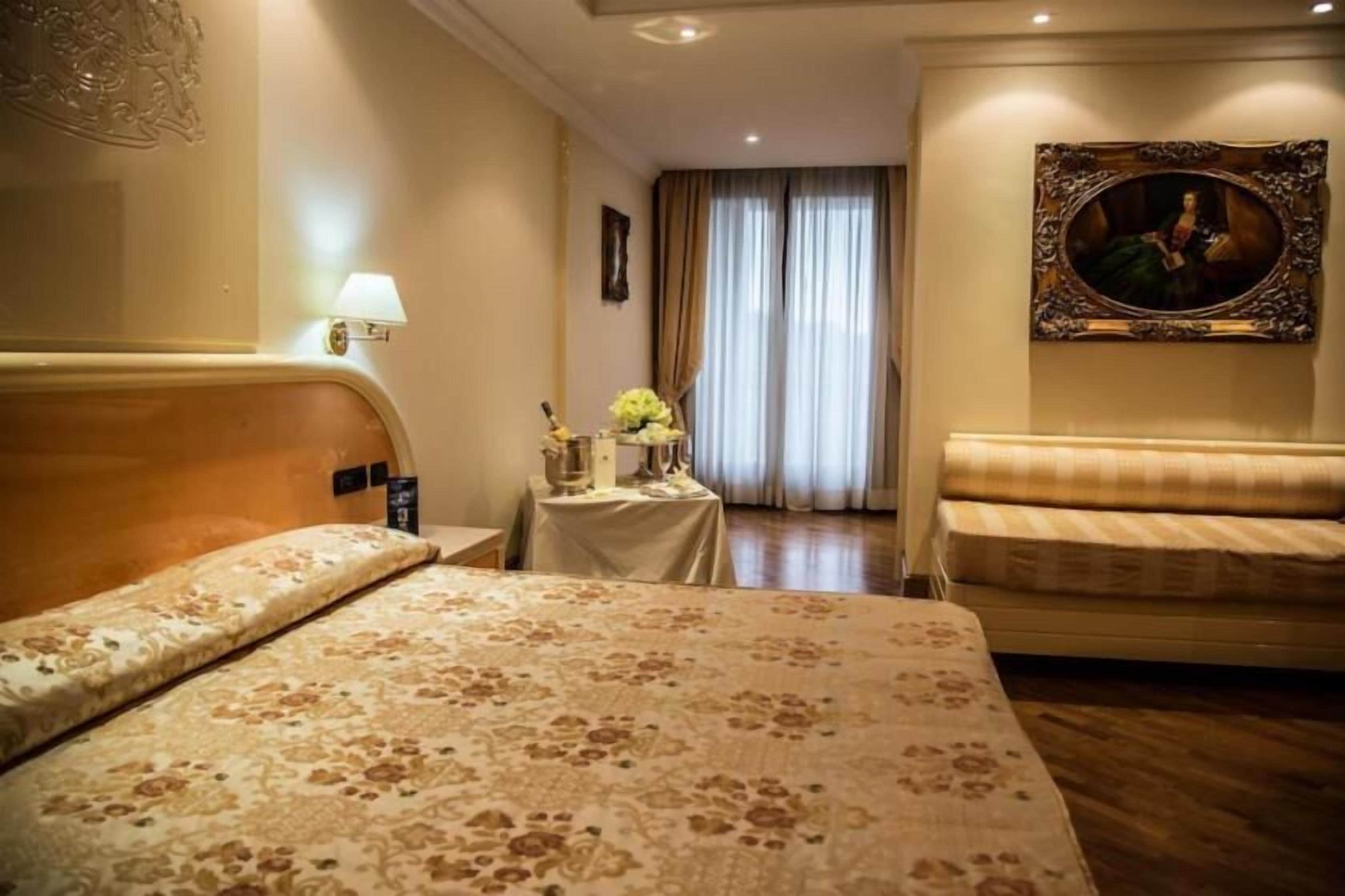A hotel room with a bed, a sofa, a small table with a lamp, and a painting on the wall.