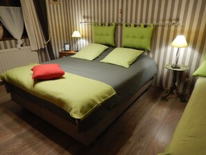 Una cama con ropa de cama verde y un almohadón rojo, una mesita de noche con una lámpara y una pared a rayas.