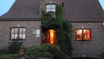 Een herenhuis van baksteen met een betegeld dak, een voordeur die van binnenuit verlicht is, en een kleine tuin met verschillende planten en bloemen.