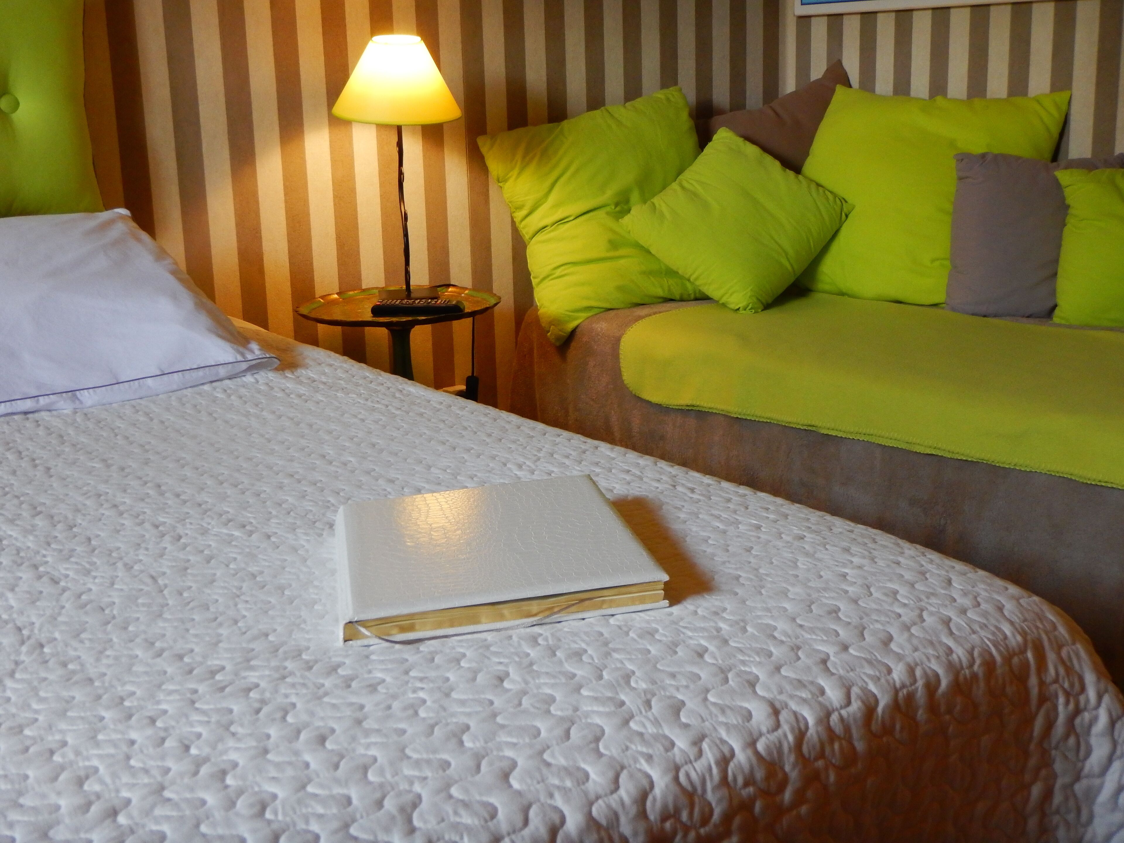 Een keurig opgemaakt bed met een witte, gestructureerde deken, een boek erop, en een groene bank met meerdere kussens op de achtergrond.