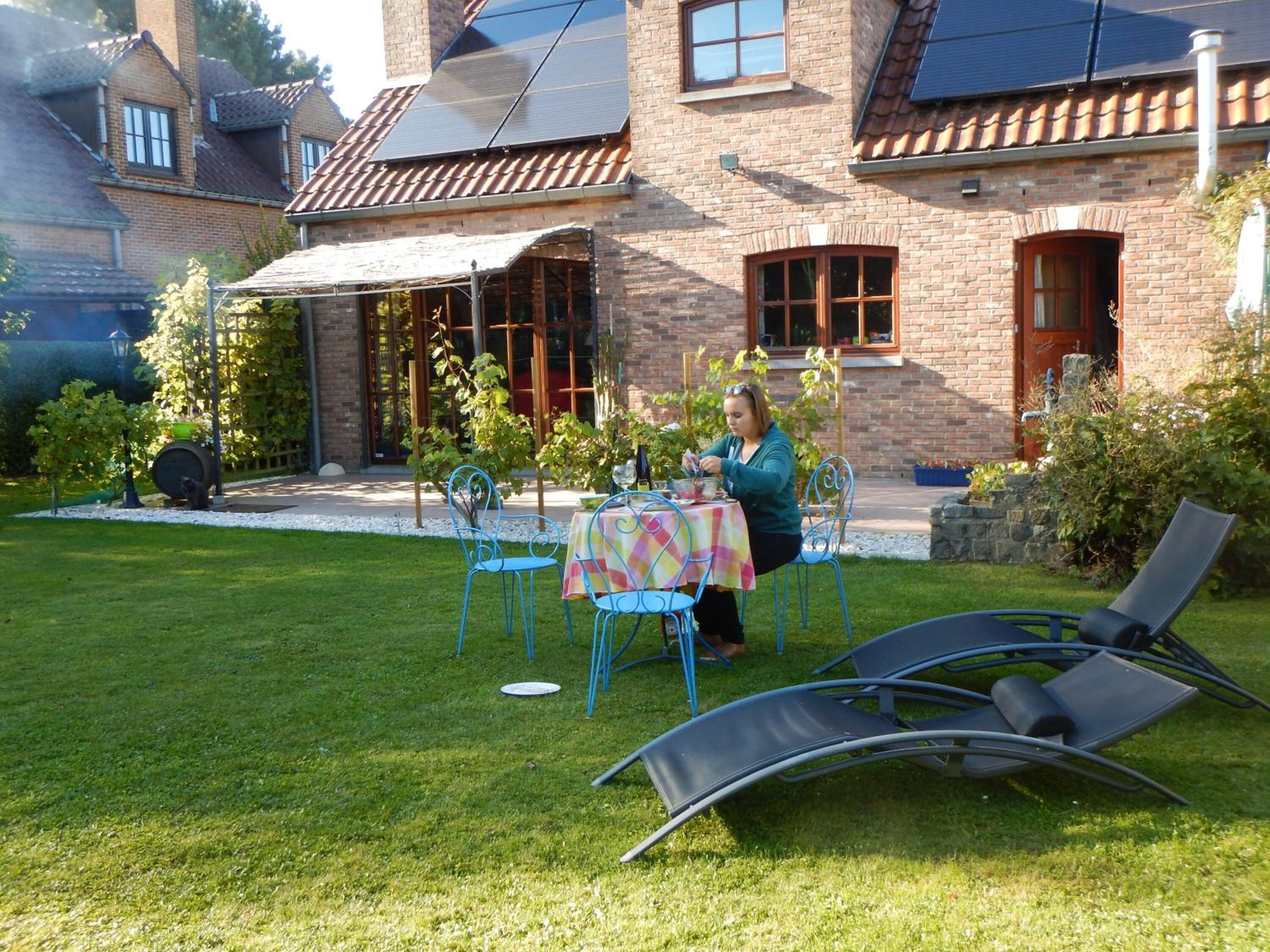 Une terrasse avec une table et des chaises, une personne assise, et un bâtiment avec des murs en briques et des fenêtres en arrière-plan.