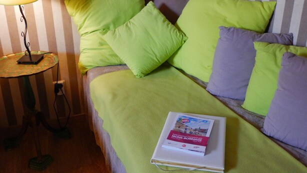 Una cama con almohadas verdes y moradas, una lámpara en una mesita de noche y un libro sobre la cama.