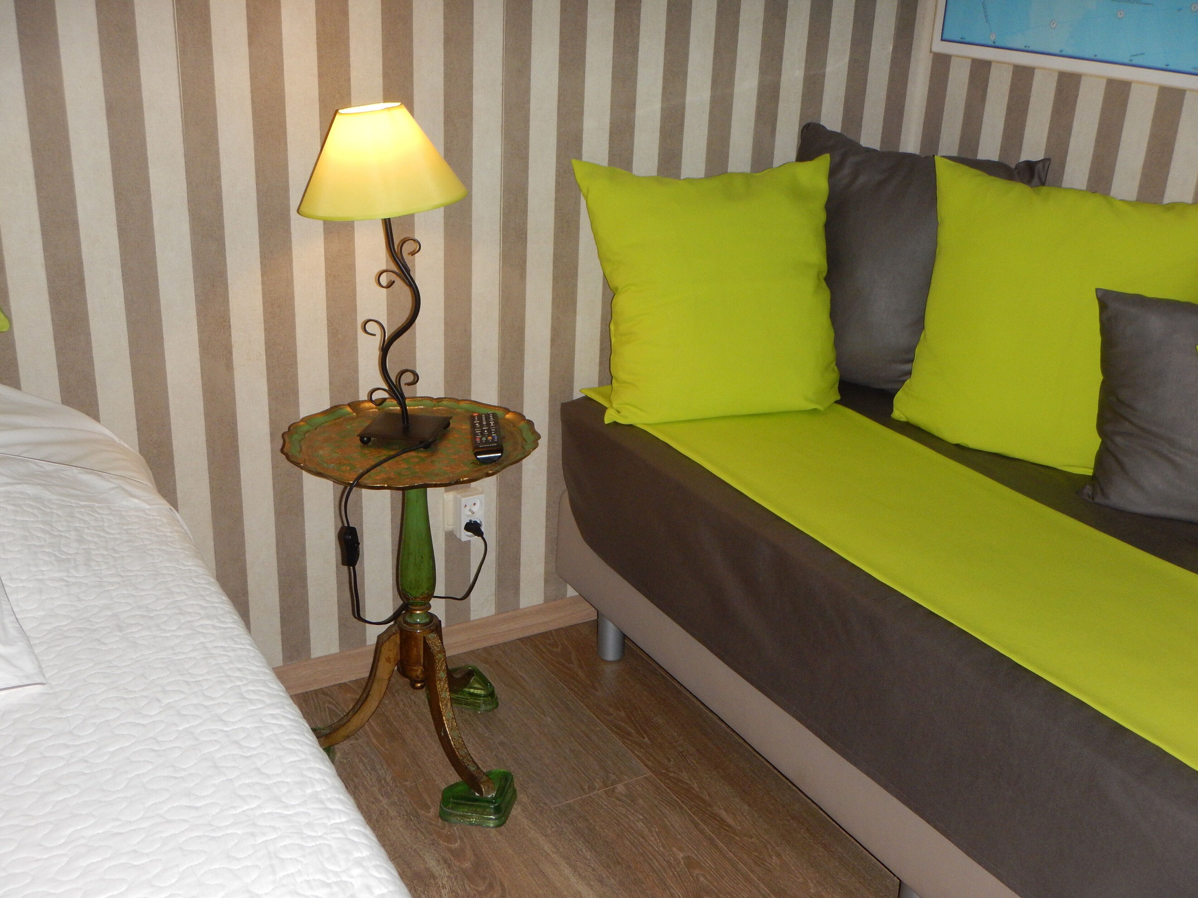 A hotel room with a striped wall, a green floor lamp, a brown sofa with green and gray pillows, and a white bed.