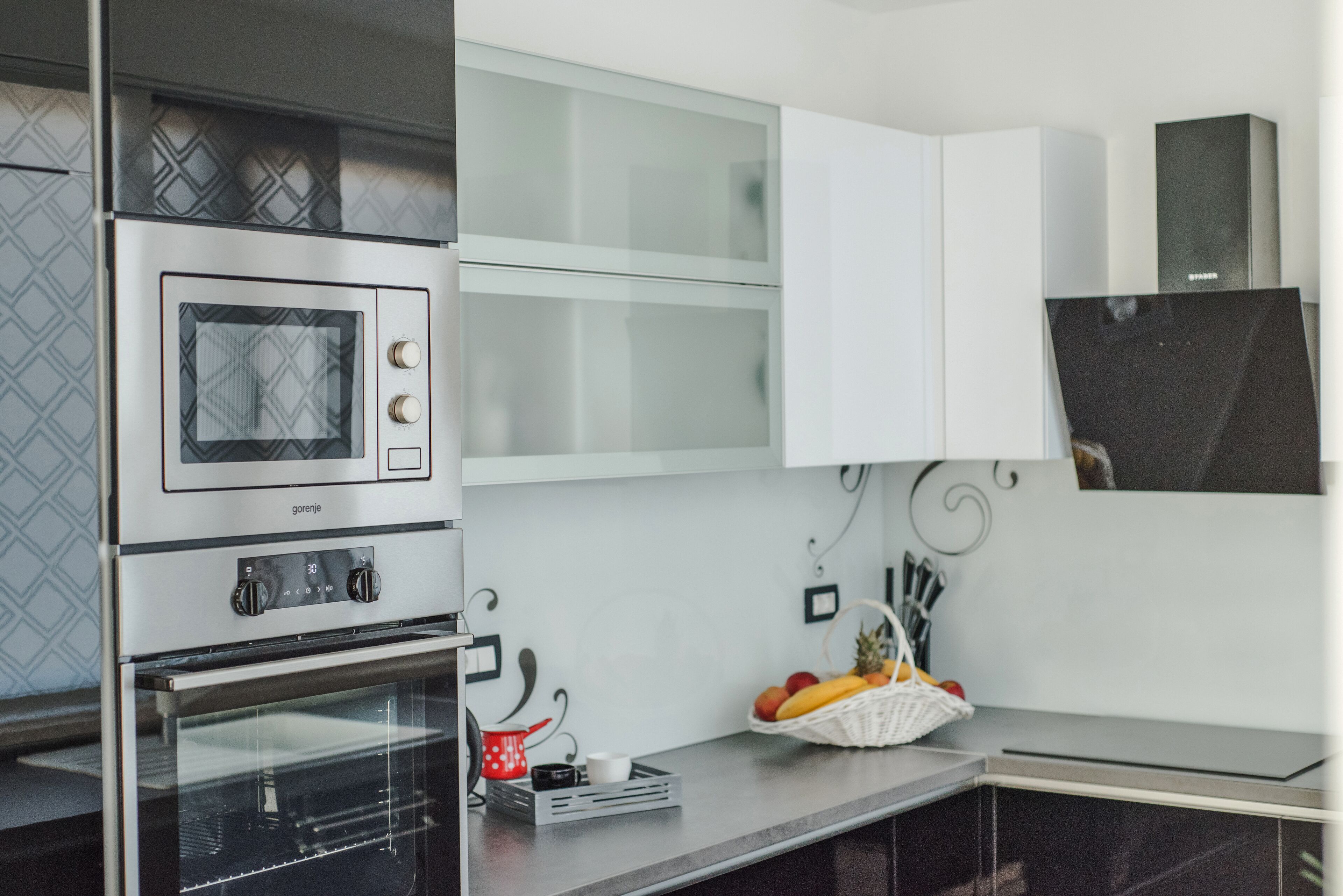 A kitchen with a built-in oven, a microwave, and a refrigerator. There is a basket of fruit on the counter.