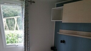 A single bed with white bedding and a yellow blanket, a window with a curtain, and a blue wall with a shelf.