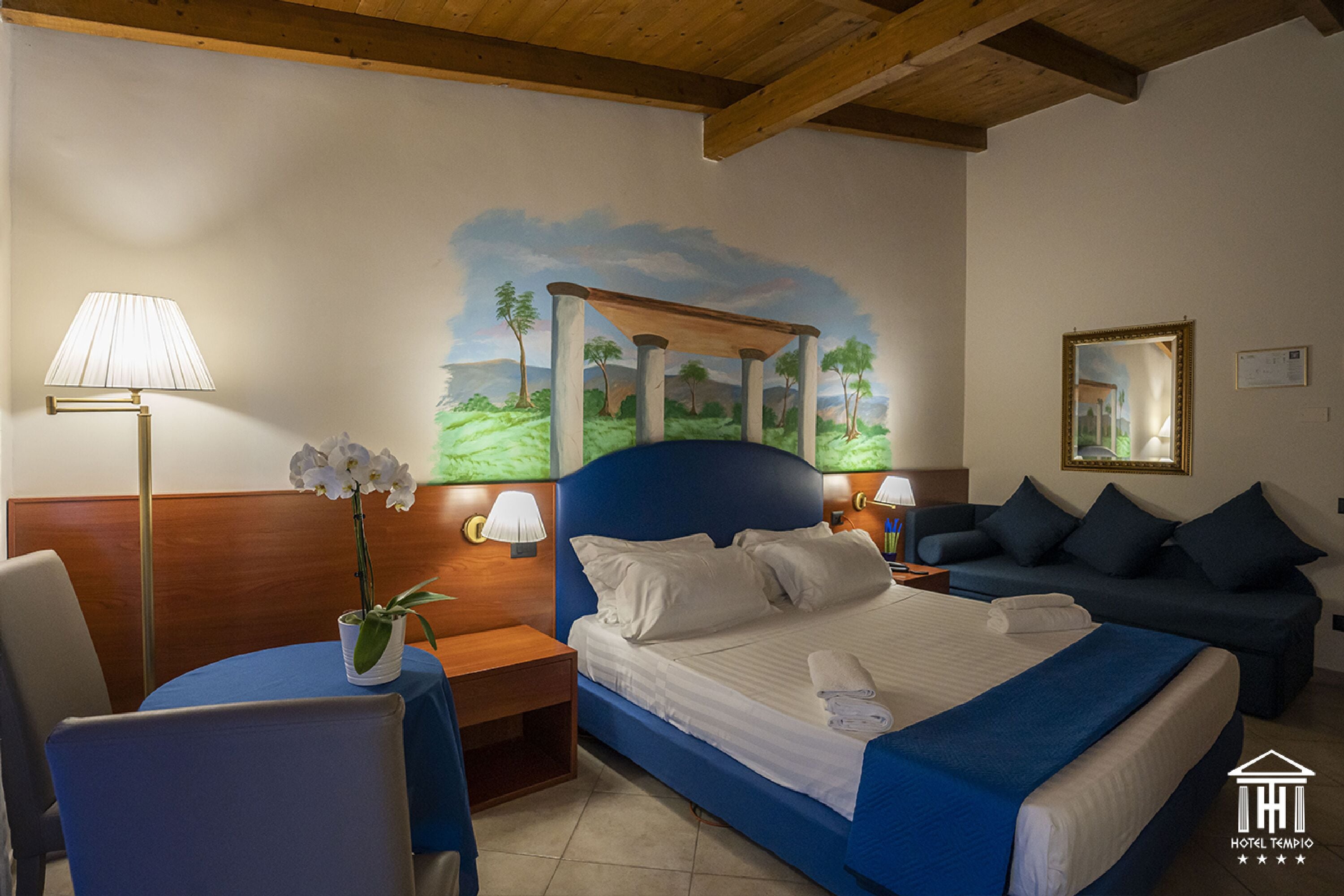 A hotel room with a bed, a blue sofa, a desk with a lamp, a chair, and a wall painting.
